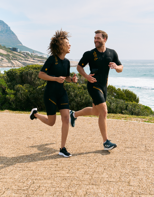 Zwei Menschen in EMS-Anzügen laufen am Strand, lächeln und genießen die Freiheit und Flexibilität ihres EMS-Trainings.