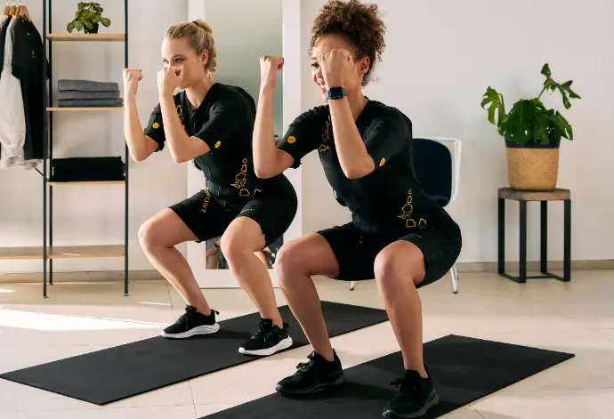 Two women in EMS suits perform squats indoors, using EMS training to strengthen their legs and build muscle efficiently.